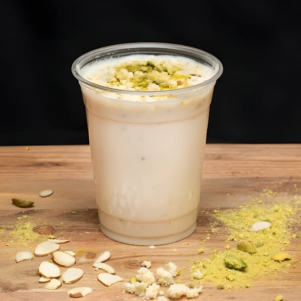 A white drink in a clear plastic cup, topped with green pistachio powder and nuts, on a wooden surface.