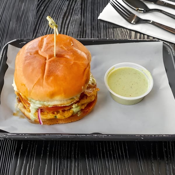 Halal chicken burger with sauce, tomatoes, and onions, served with a side of green sauce.