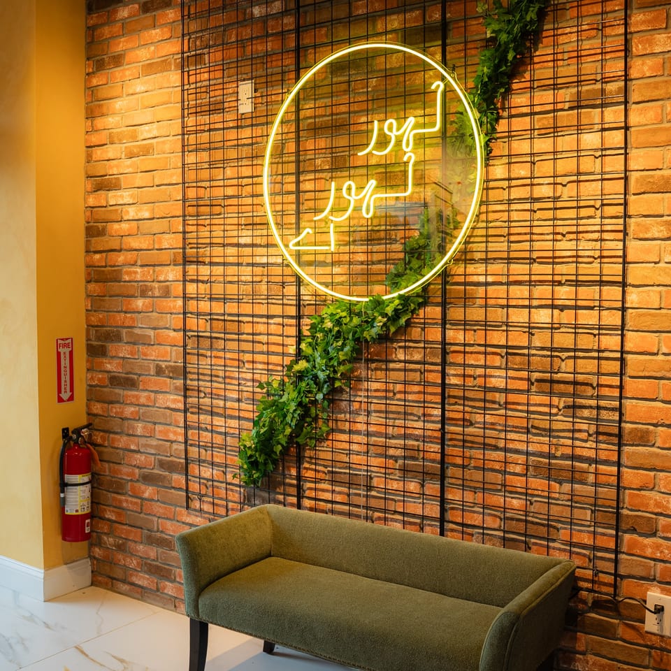 A green bench sits against a brick wall with a neon sign and green vine on a black grid.