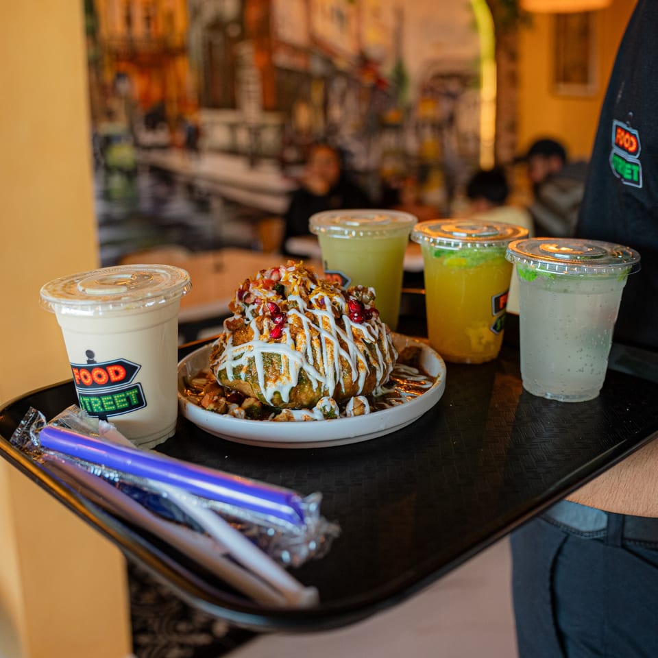 A server carries a tray with a large chaat dish and four drinks from Food Street restaurant.