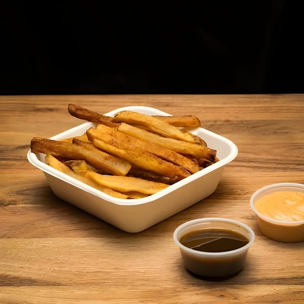 A white container of golden-brown fries sits on a wooden table with two dipping sauces.