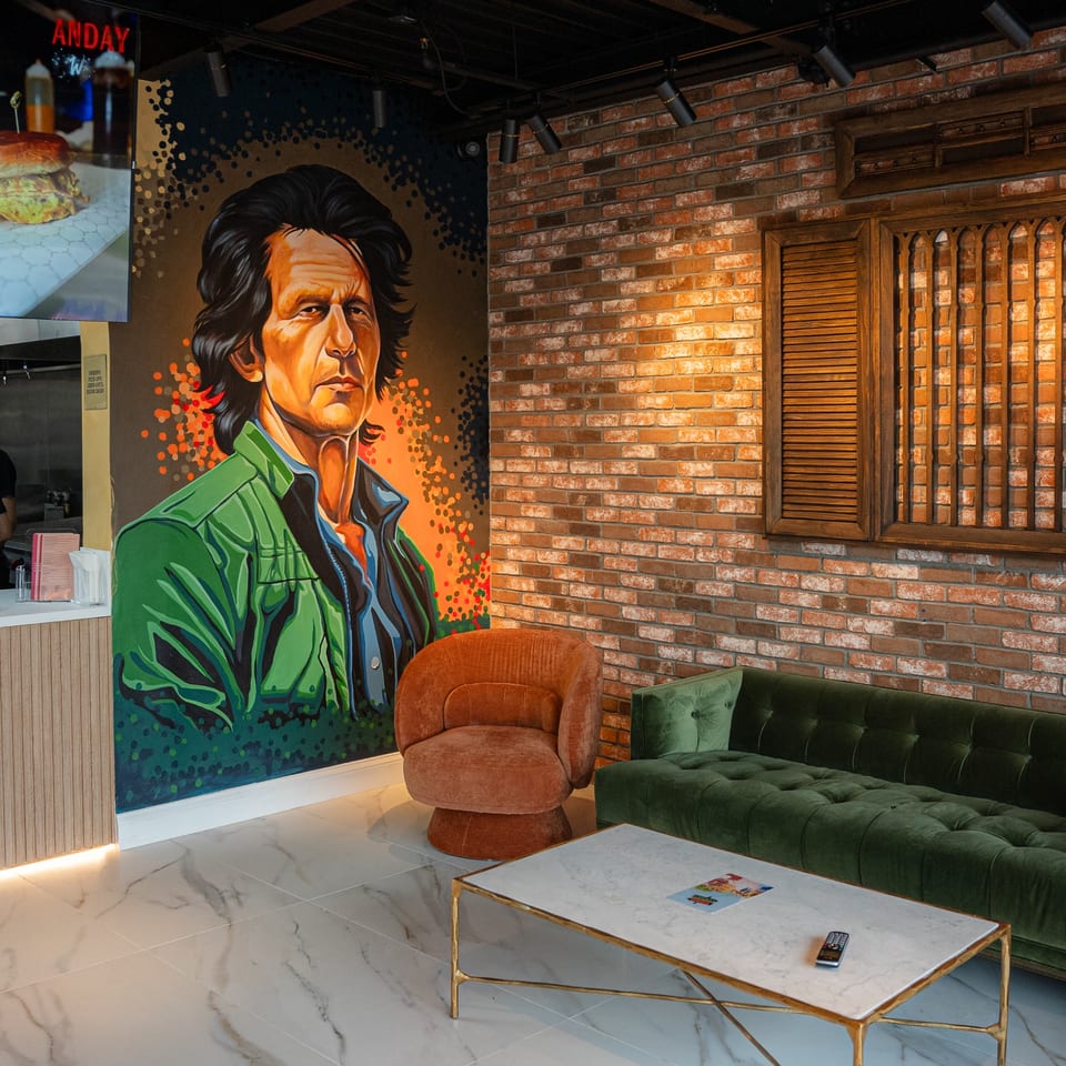 Restaurant interior with a large mural, brick wall, green sofa, orange chair, and marble table.