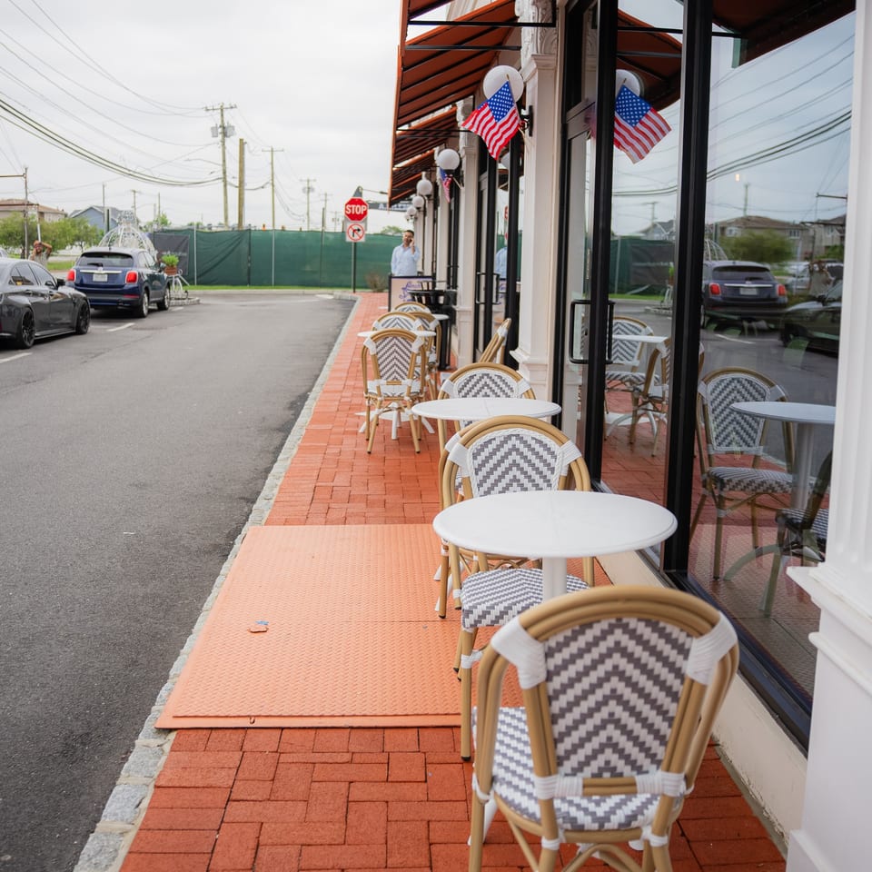Outdoor dining area with white tables and woven chairs on a red brick sidewalk, American flags adorn the building.